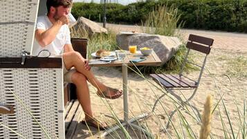 A beachside setting with a wicker lounge chair, a table with breakfast items, and a metal lamp post.