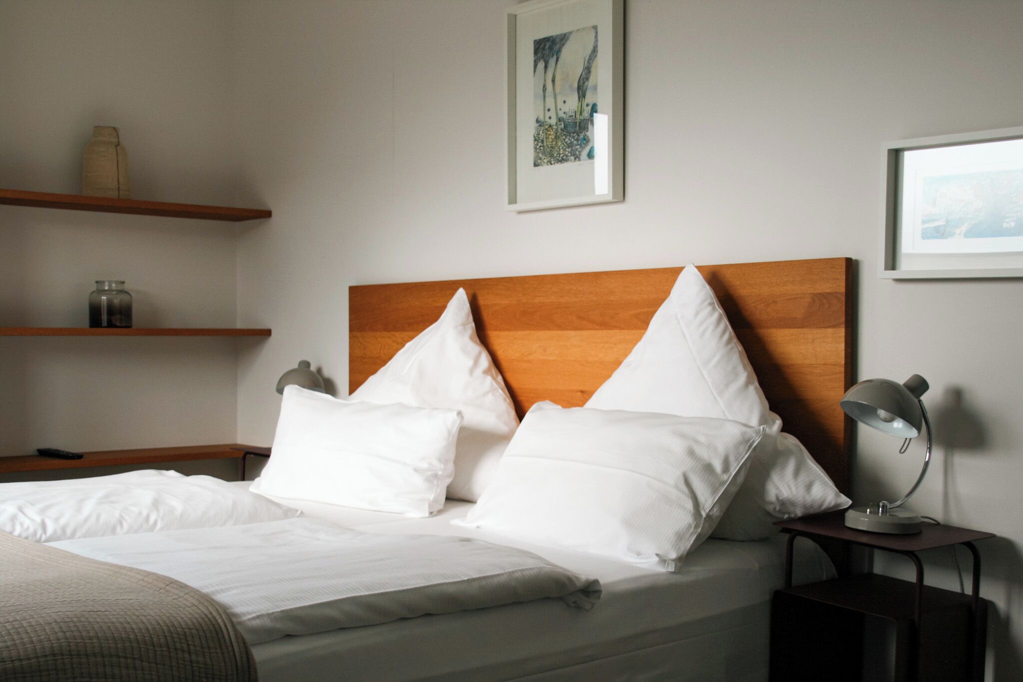 Een net opgemaakt bed met witte lakens en kussens, een houten hoofdbord, een nachtkastje met een lamp en twee planken met decoratieve objecten.