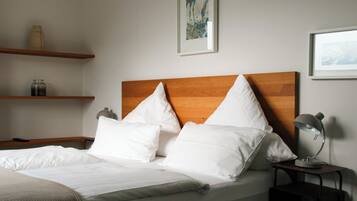 Een net opgemaakt bed met witte lakens en kussens, een houten hoofdbord, een nachtkastje met een lamp en twee planken met decoratieve objecten.