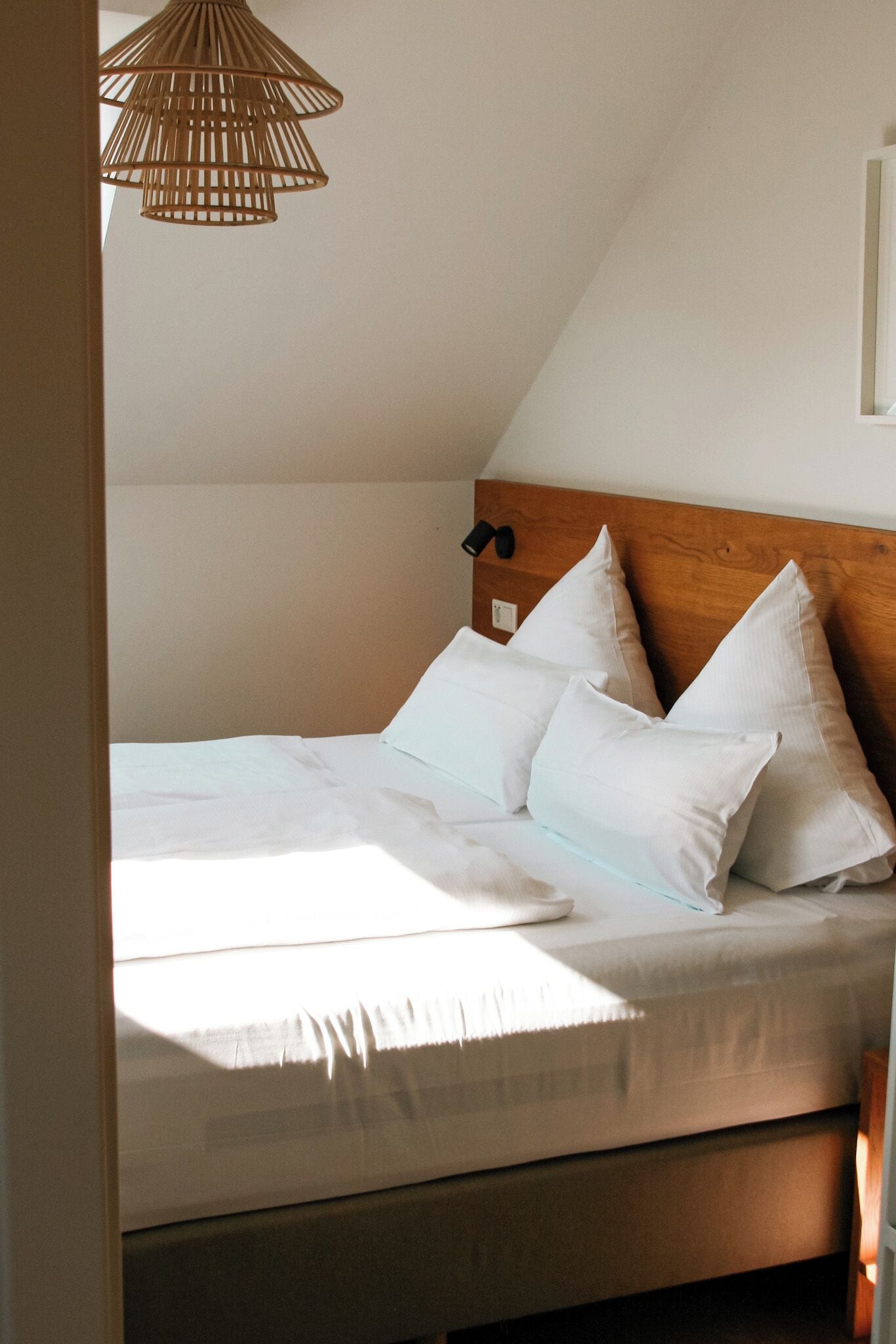 A neatly made bed with white linens and pillows, a wooden headboard, and a woven light fixture above.