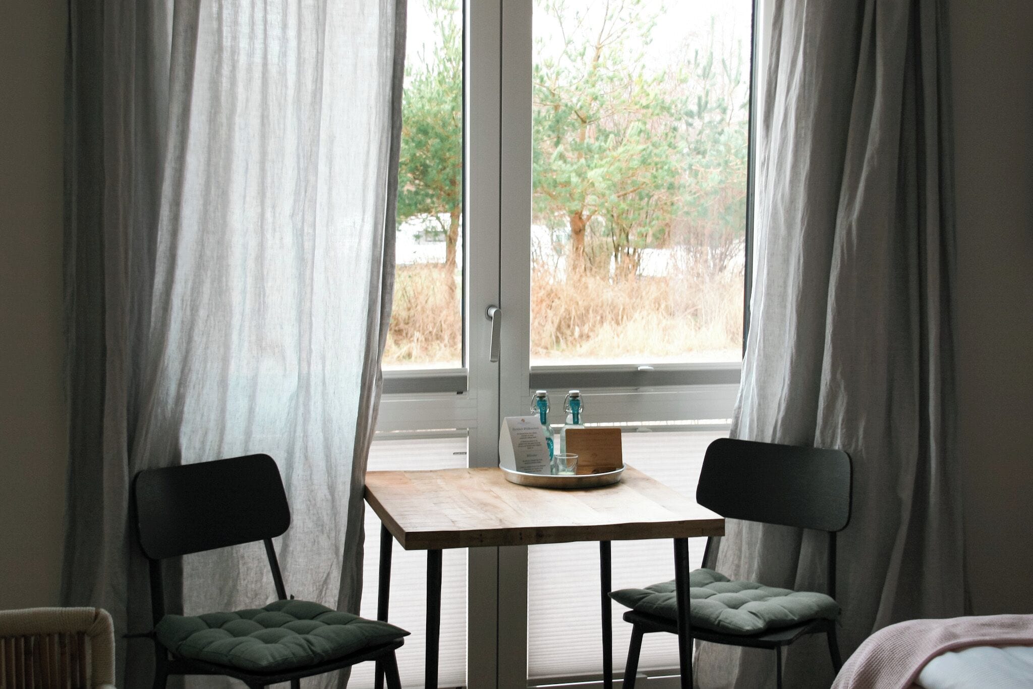 Una habitación con una mesa y dos sillas, una ventana con cortinas, y vistas a árboles fuera.