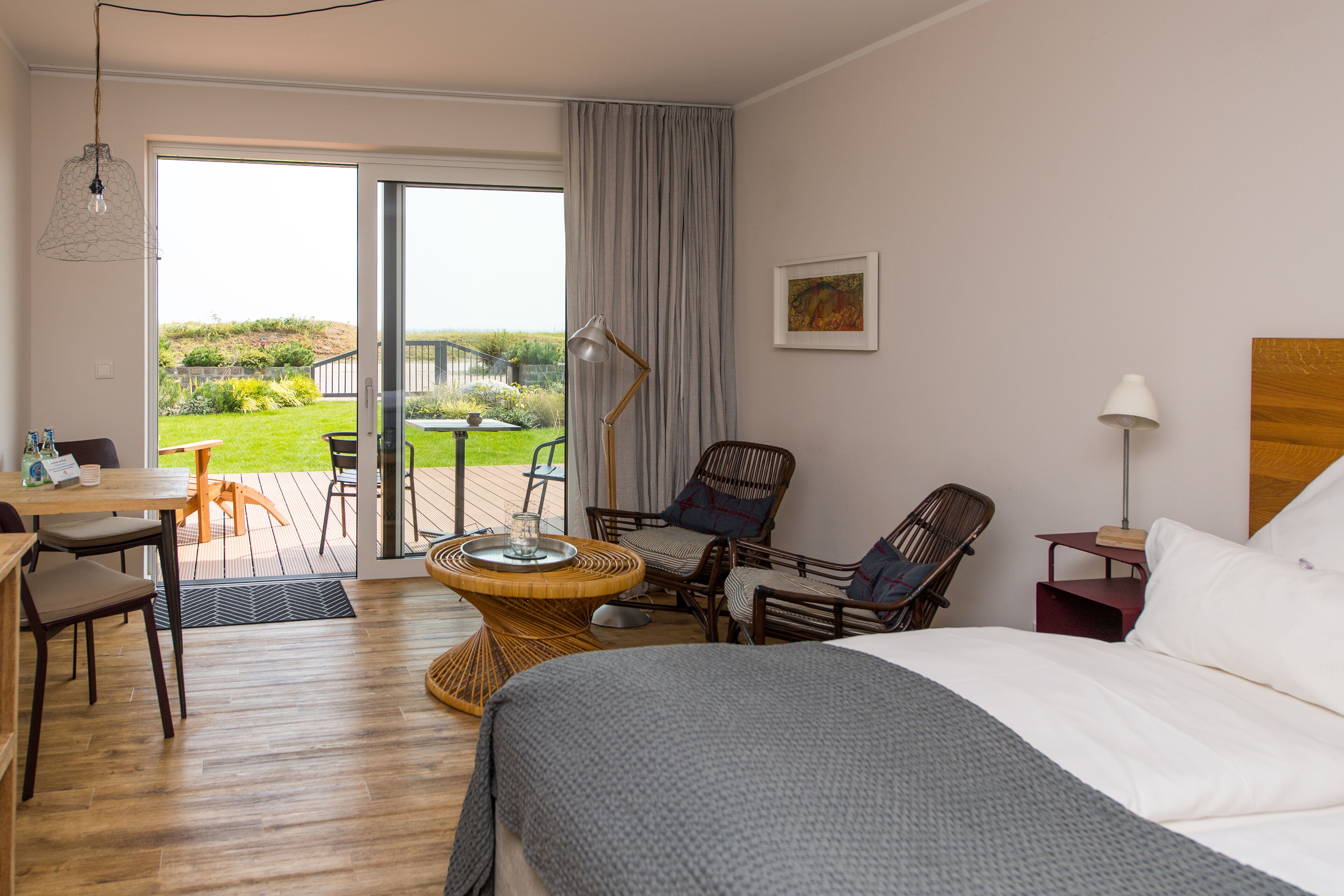 A hotel room with a bed, a dining table, chairs, a wicker table, a lamp, and a view of the outdoors.