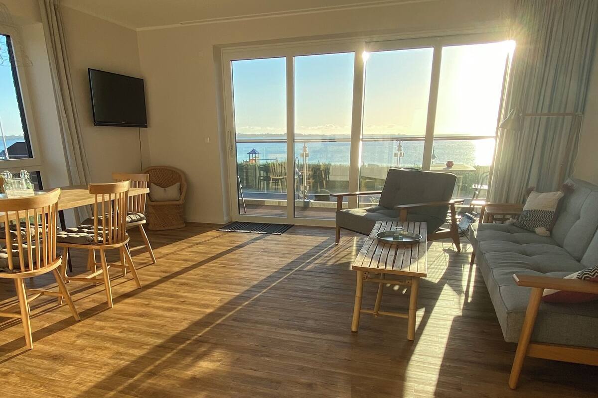 Een ruime woonkamer met houten vloer, een bank, een eettafel met stoelen en een groot raam met uitzicht op zee.