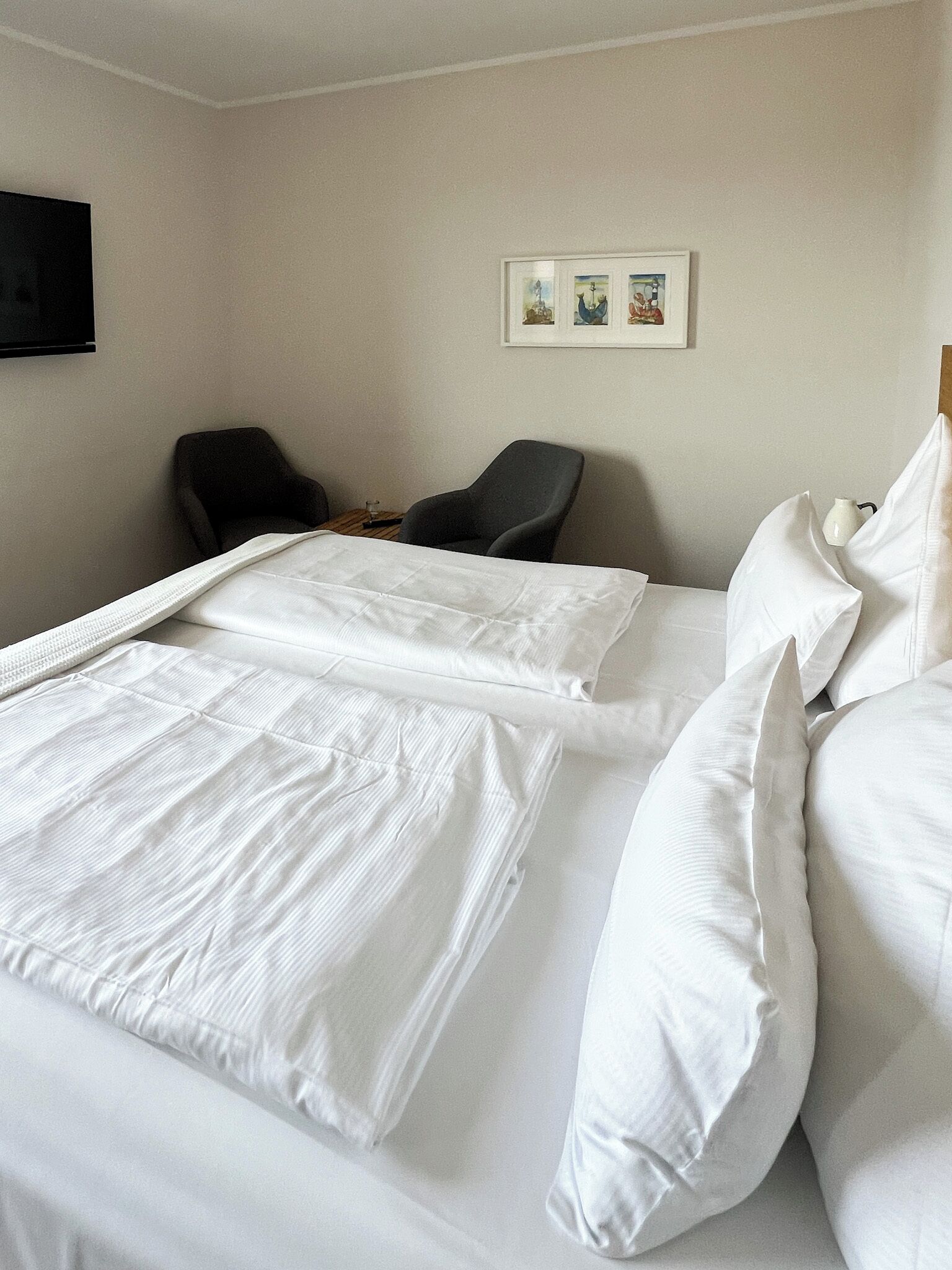 Ett hotellrum med en snyggt bäddad säng, två stolar, en platt-TV som är monterad på väggen och ett litet bord med en vas.