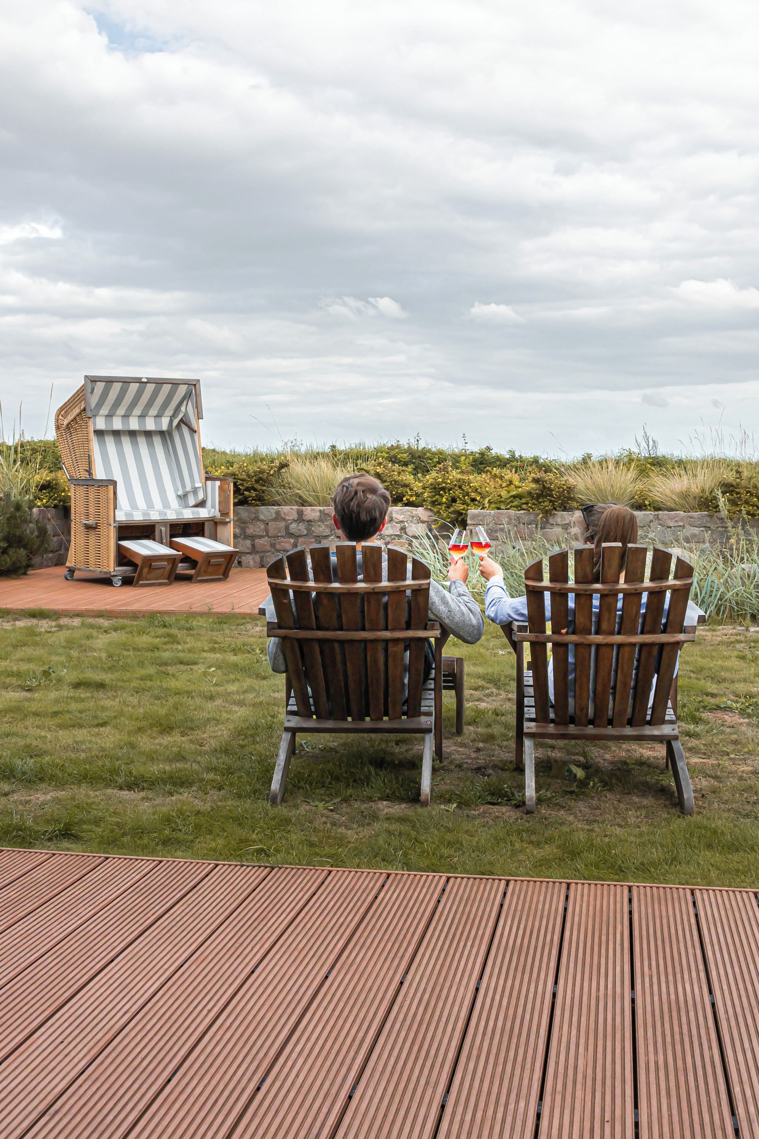 Two wooden chairs facing a wicker lounge chair and a stone wall.