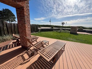 A wooden deck with outdoor furniture, a brick pillar, and a view of a grassy area with a fence and a street lamp in the background.