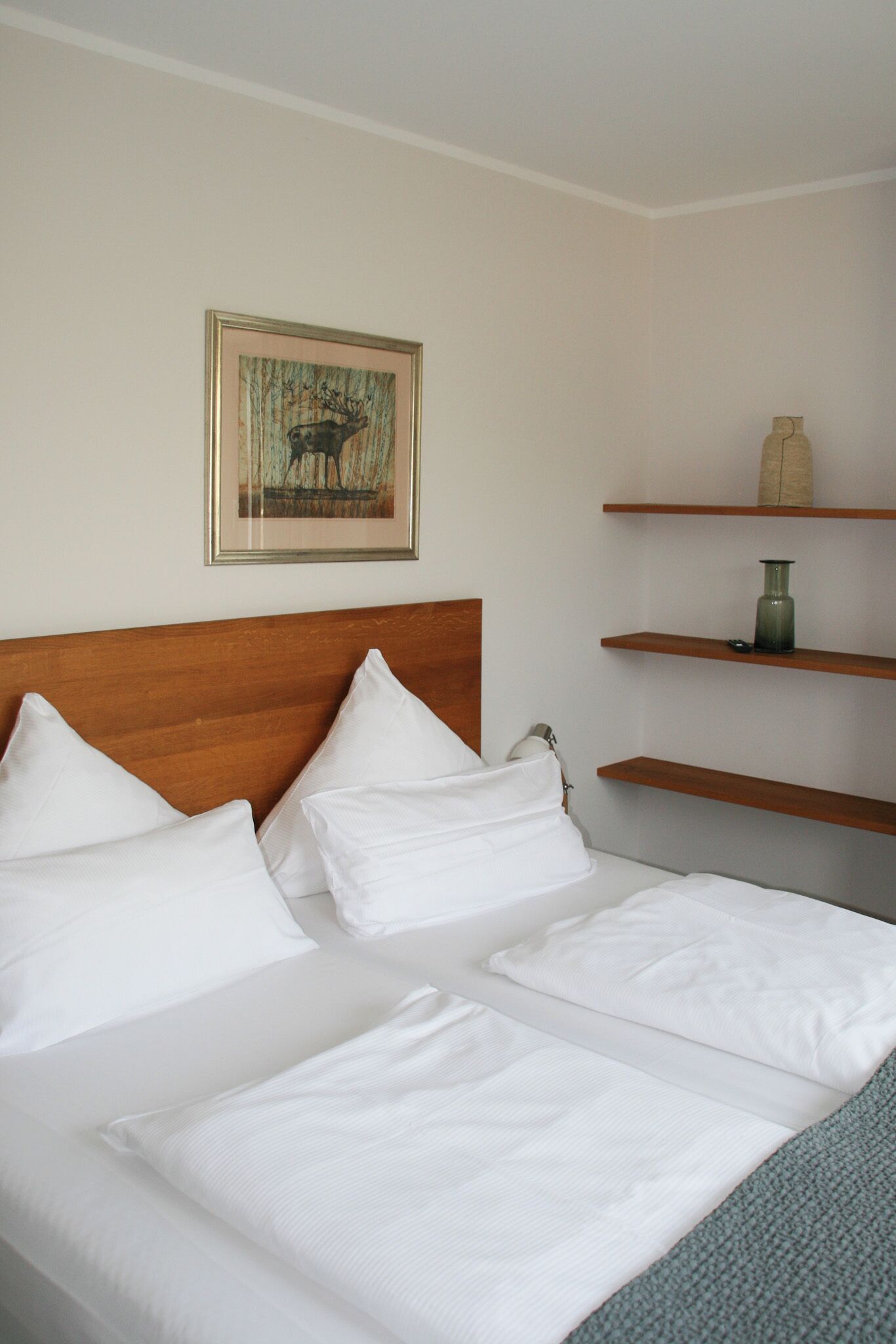 Una cama bien hecha con ropa blanca y cabecera de madera. Encima de la cama hay un cuadro enmarcado en la pared y una estantería con un jarrón y un objeto pequeño.