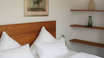 Una cama bien hecha con ropa blanca y cabecera de madera. Encima de la cama hay un cuadro enmarcado en la pared y una estantería con un jarrón y un objeto pequeño.