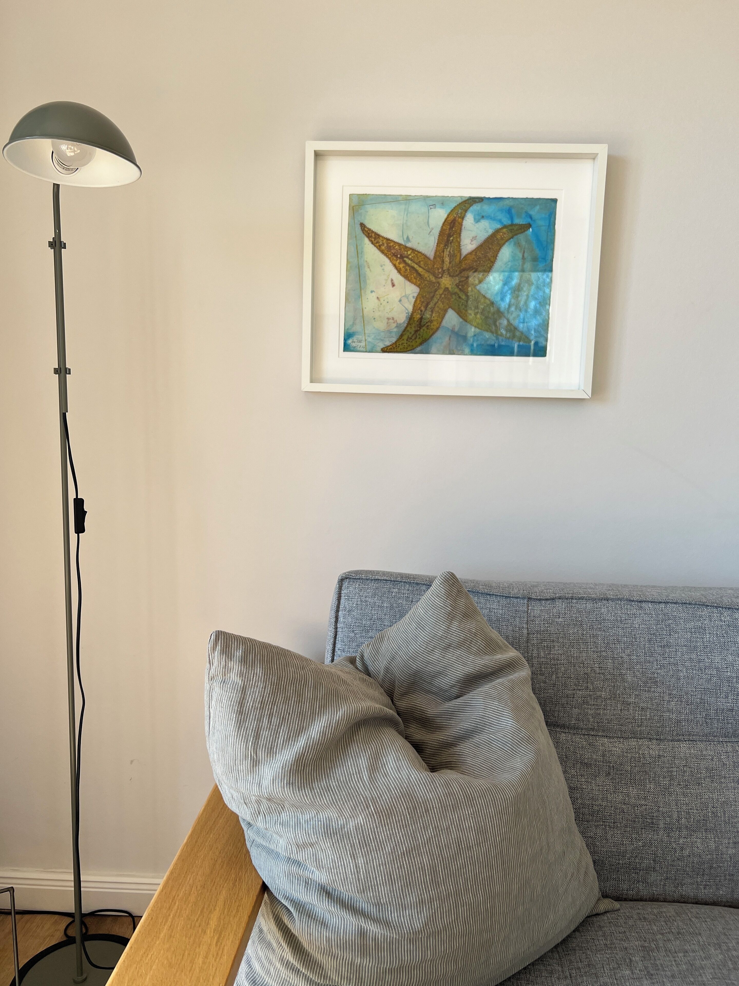 Una obra d'art amb un marc que mostra una estrella de mar a la paret, un sofà gris amb coixins ratllats i una làmpada de terra amb pantalla blanca.