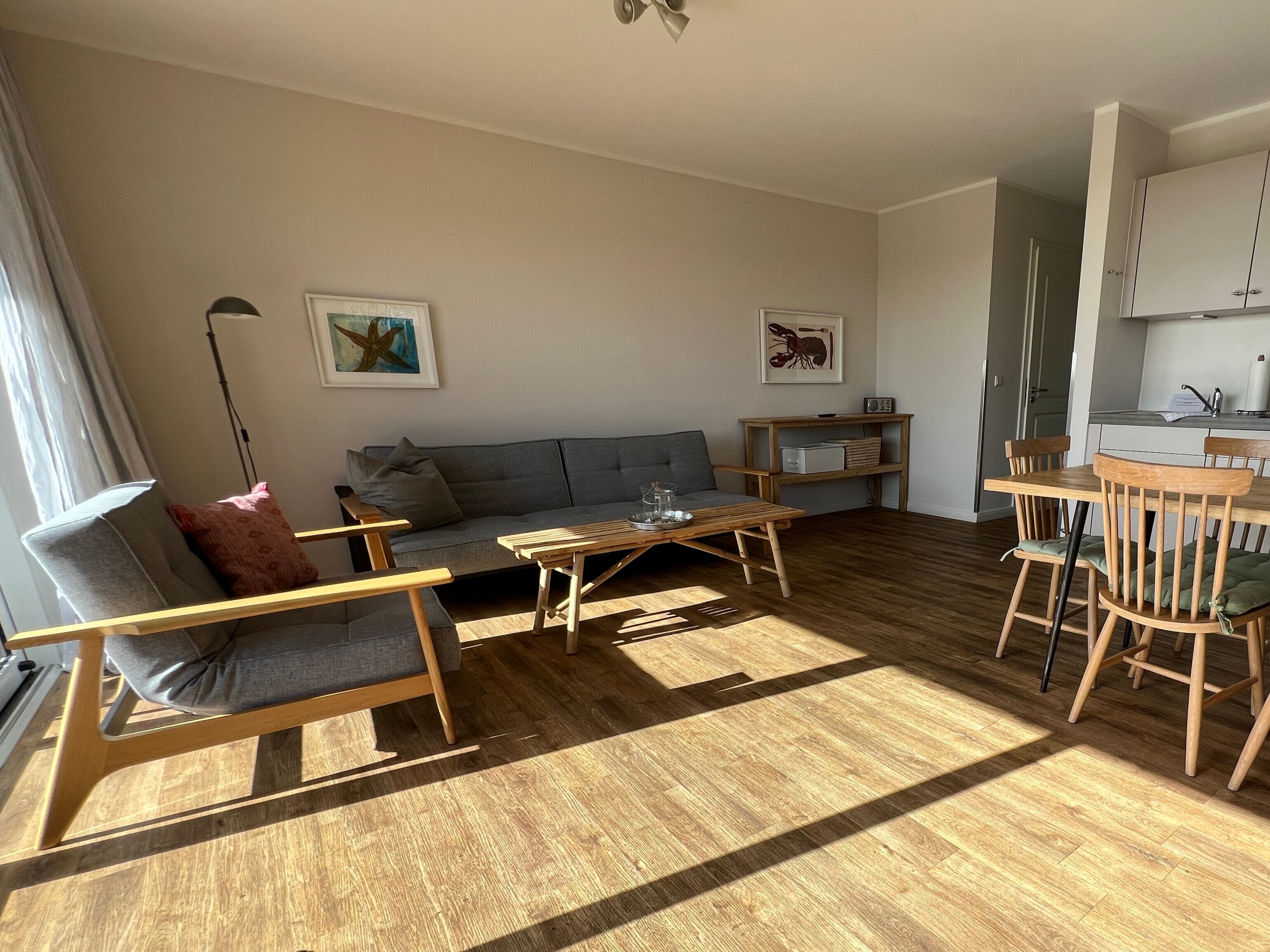 A modern living room with a grey sofa, wooden chairs, a coffee table, and a dining area with a table and chairs. There are two framed pictures on the wall, a lamp, and a kitchen area in the background.