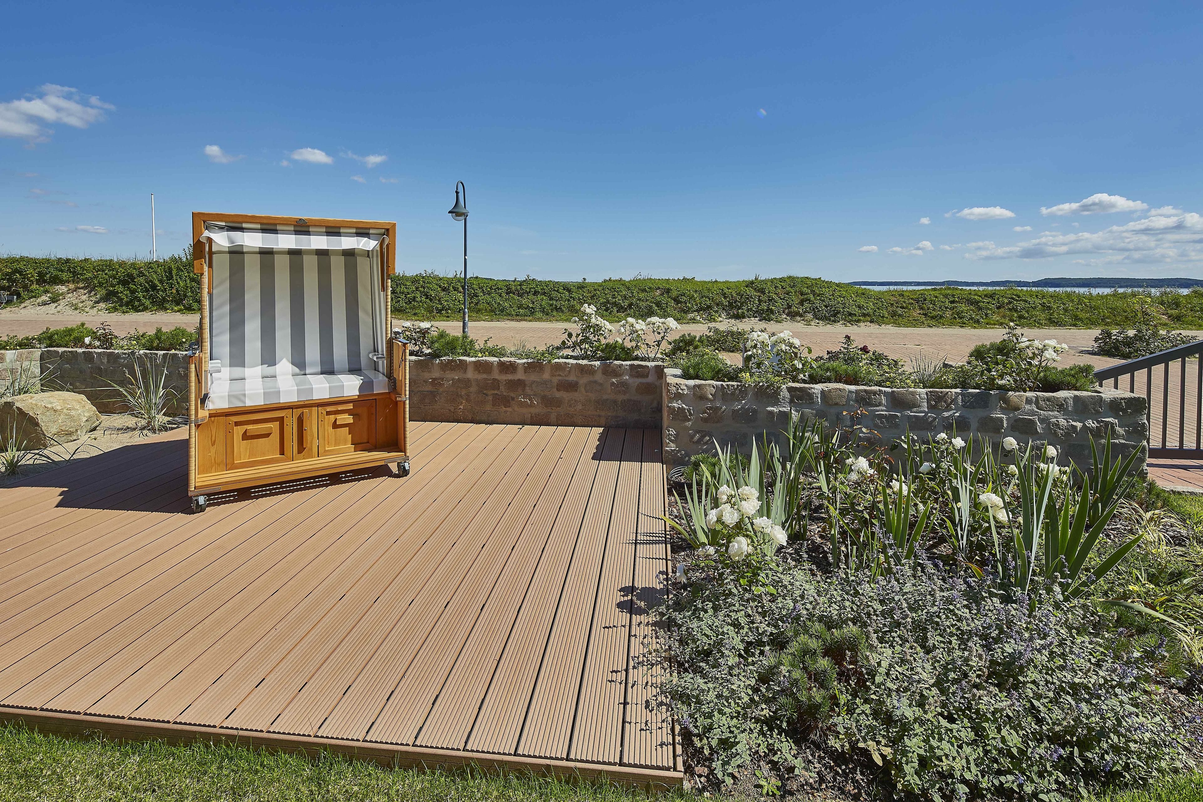 A wooden deck with a striped beach chair, surrounded by greenery and a view of the beach.