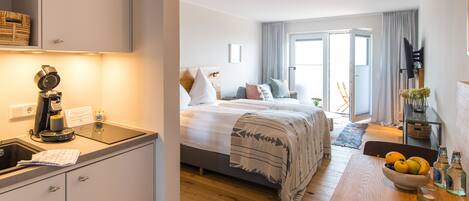 Una habitación de hotel moderna con suelo de madera, una cama con manta estampada, una mesa de comedor con frutero y una kitchenette con cafetera.