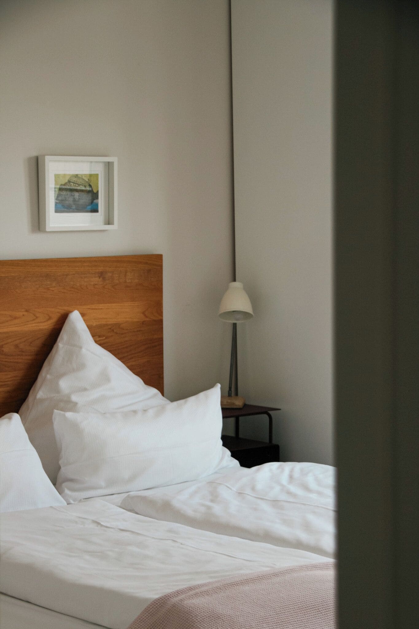 Un lit bien fait, avec du linge de lit blanc, une tête de lit en bois et une petite table de chevet sur laquelle se trouve une lampe.