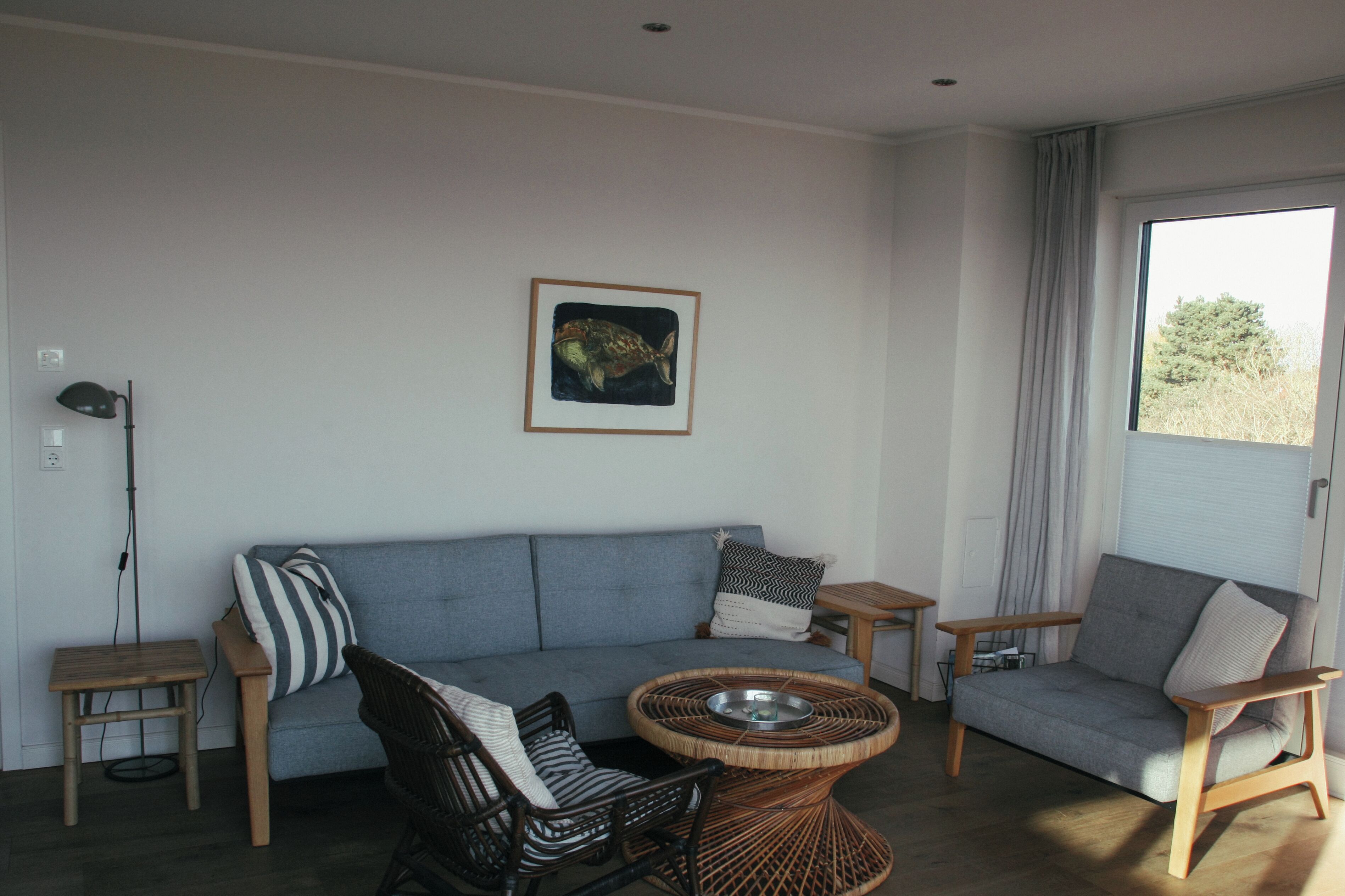 Una sala de estar con un sofá gris, dos sillones, una mesa de centro de mimbre y un cuadro en la pared.