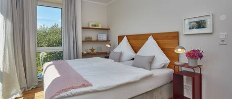 A neatly made bed with a wooden headboard, a bedside table with a lamp and a flower vase, and a window with curtains.