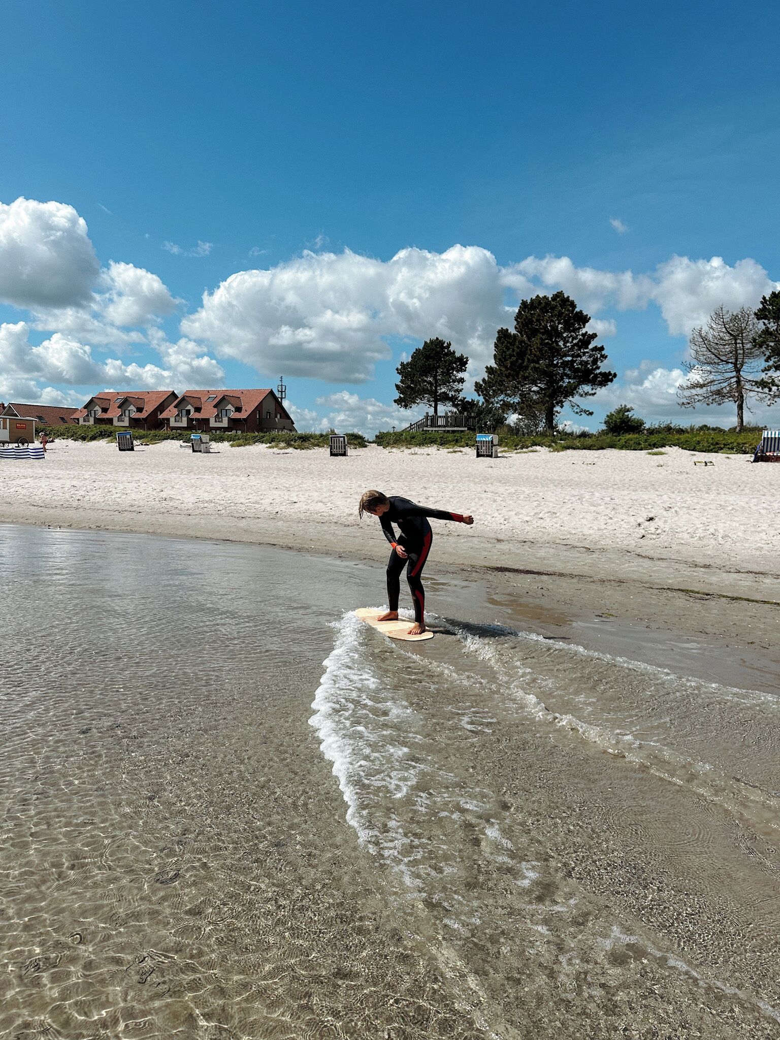 A person surfing on a beach with clear water, a sandy shore, and a row of houses in the background.