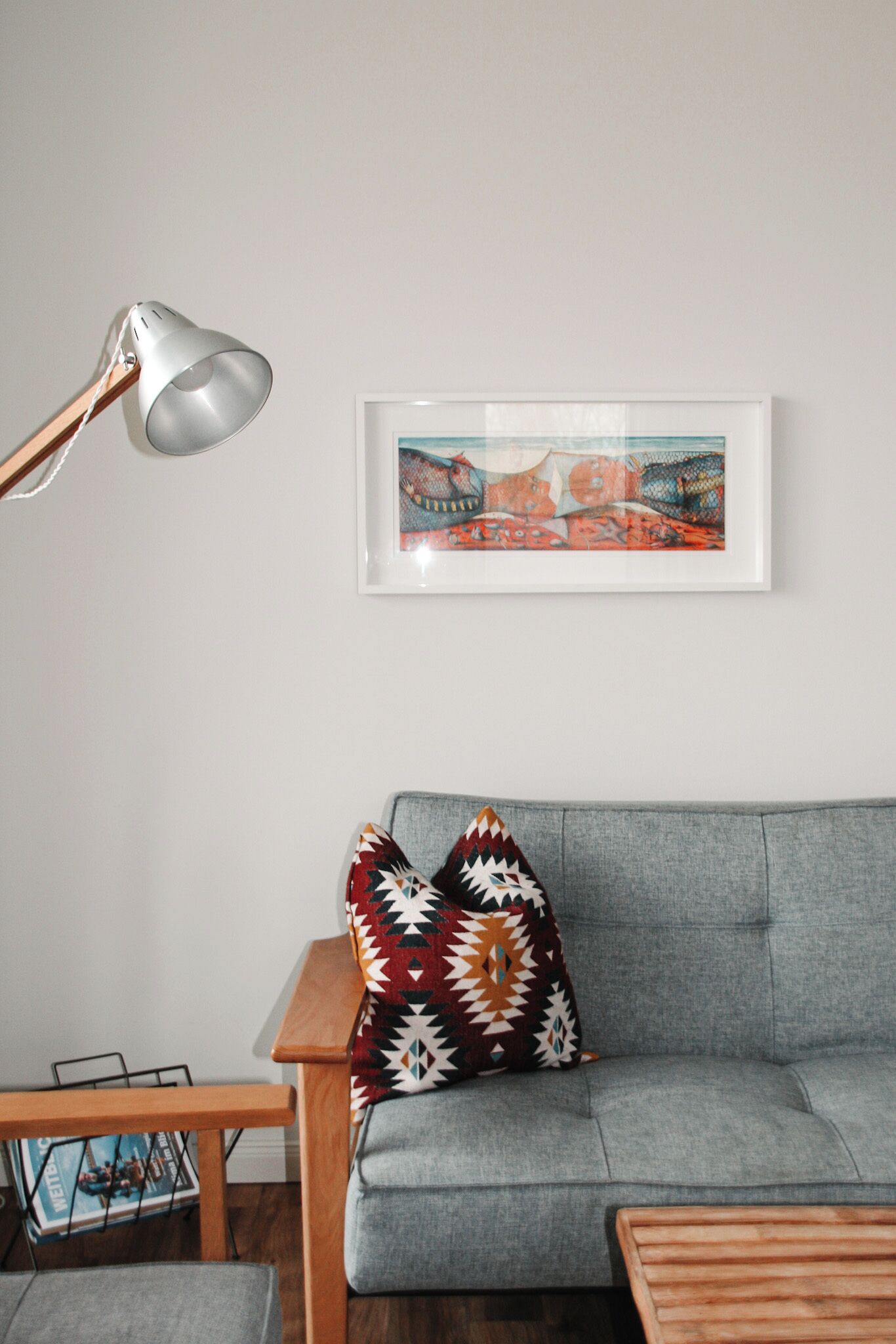 Ein modernes Wohnzimmer mit einem grauen Sofa, einem Holznachttisch, einer Stehlampe und einem farbenfrohen, abstrakten Gemälde.