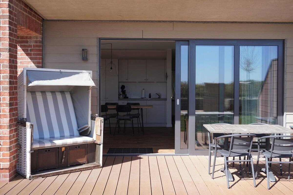 Een terras met een barbecue, een tafel en stoelen, dat uitkomt op een glazen schuifdeur naar een keuken.