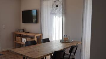 Uma sala de jantar com uma mesa de madeira, cadeiras, uma TV fixada na parede e uma janela com cortinas.