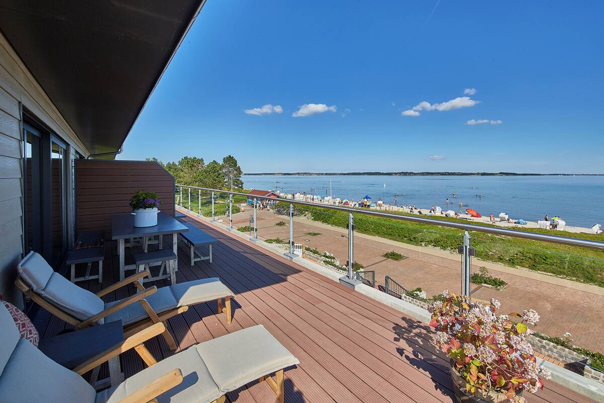 Een houten terras met lounge fauteuils en een tafel, met uitzicht op een jachthaven vol boten en een helderblauwe lucht.