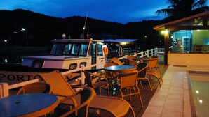 A boat docked at a waterfront restaurant with wicker chairs and tables, a palm tree, and a mountain in the background. - Pousada Porto Canal (Cabo Frio)