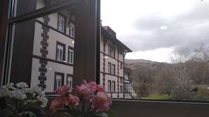 A view from a window with two potted plants, a building with traditional architecture, and a mountainous landscape. - Balneario Casa Pallotti (Valle de Carranza)