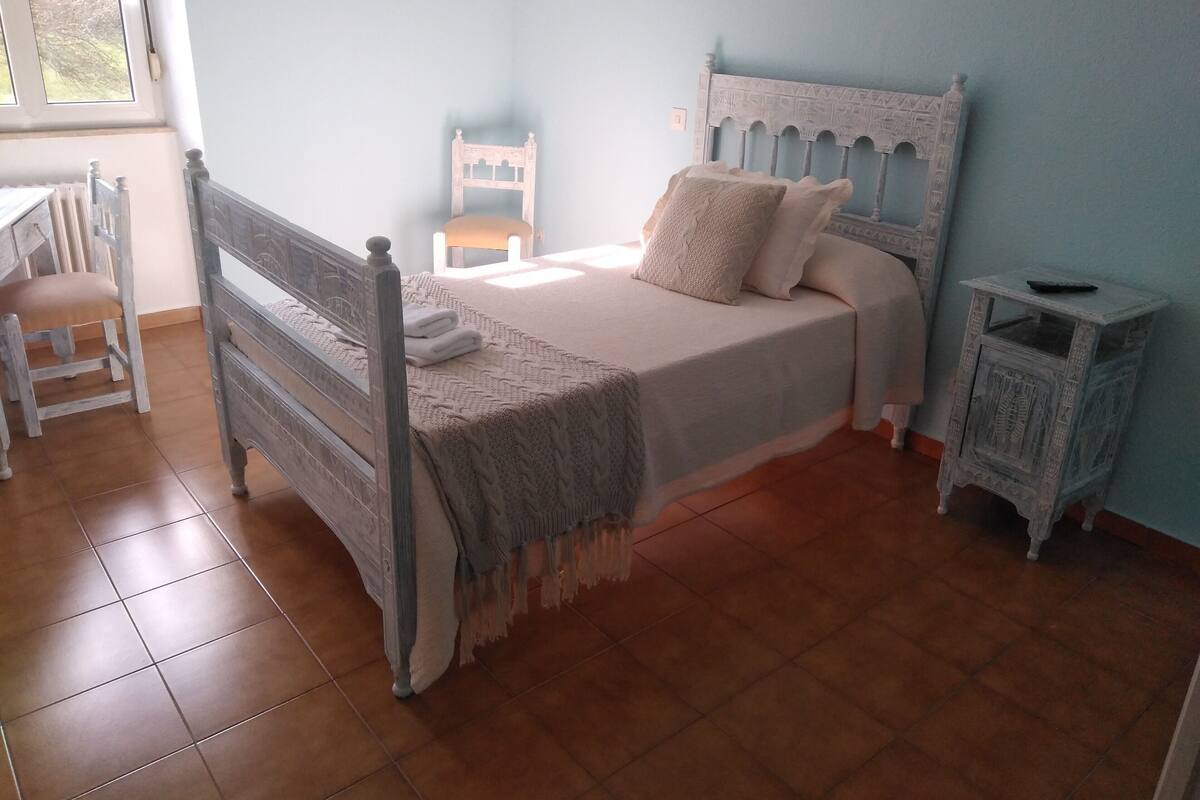 A bedroom with a bed, a chair, a nightstand, and a window.