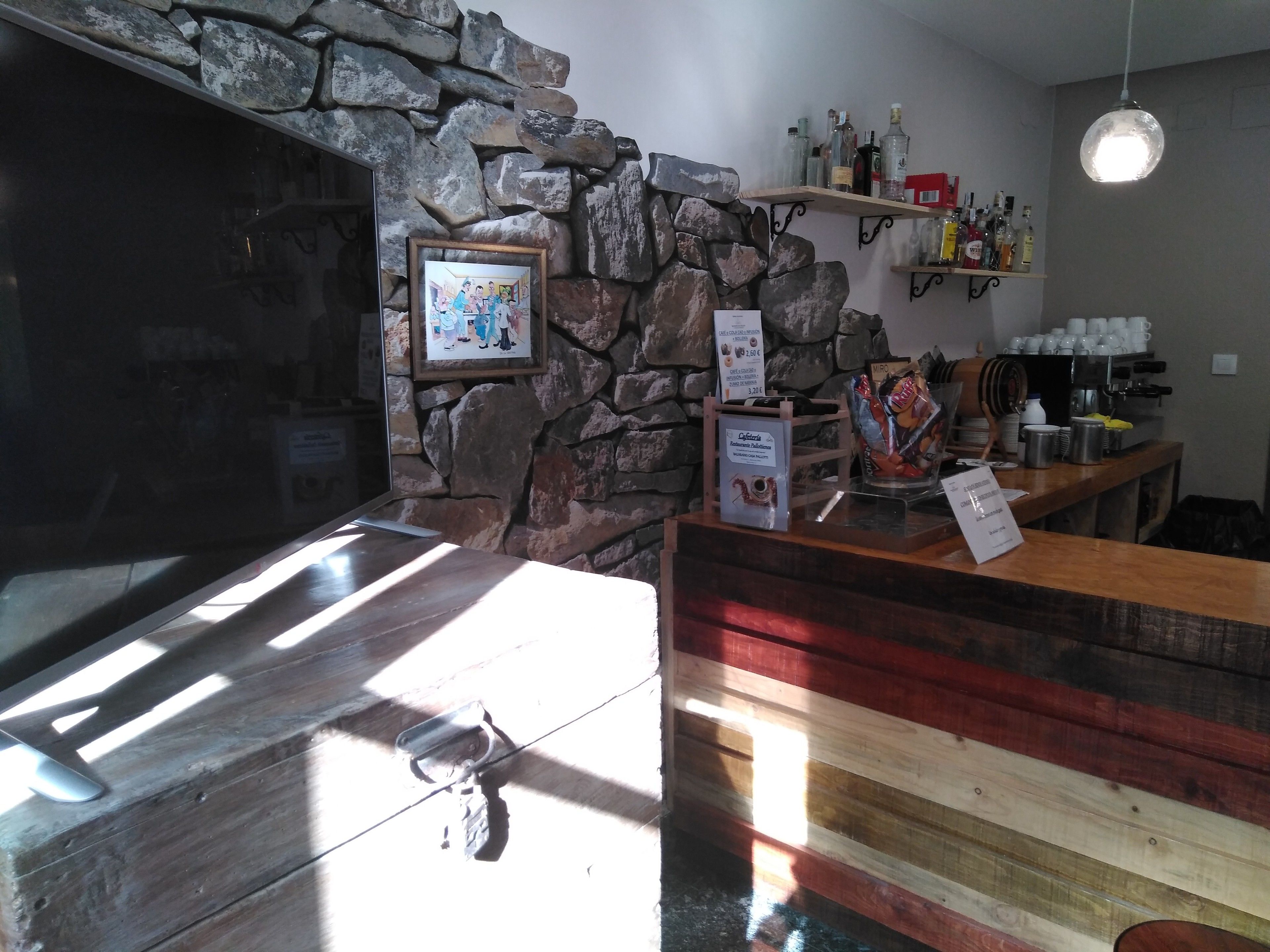 A bar area with a stone wall, wooden counter, and various bottles and cups.