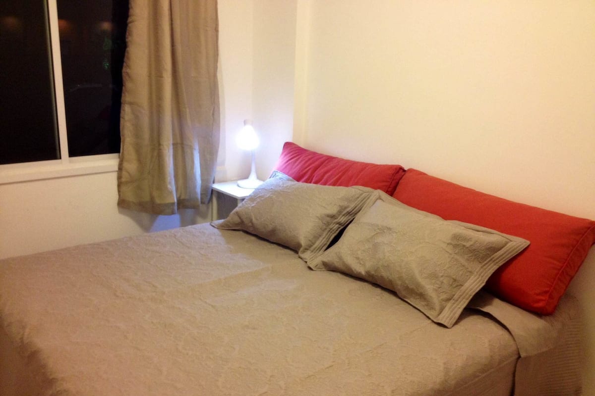 A bed with a beige bedspread and two pillows, one red and one grey, in a room with a window and a nightstand with a lamp.