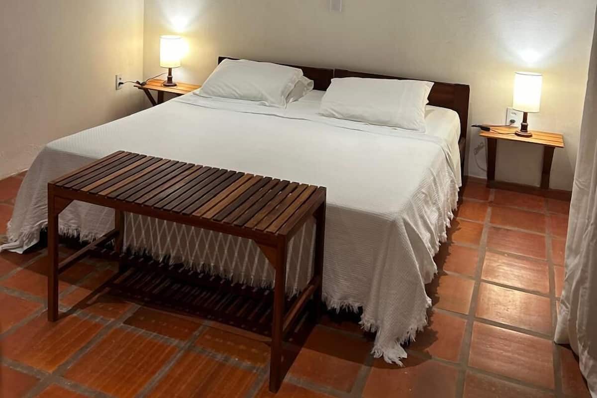 Un dormitorio con techo de madera, una cama con ropa de cama blanca, dos mesitas de noche con lámparas, un banco de madera y un reloj de pared.