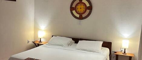 Un dormitorio con techo de madera, una cama con ropa de cama blanca, dos mesitas de noche con lámparas, un banco de madera y un reloj de pared.