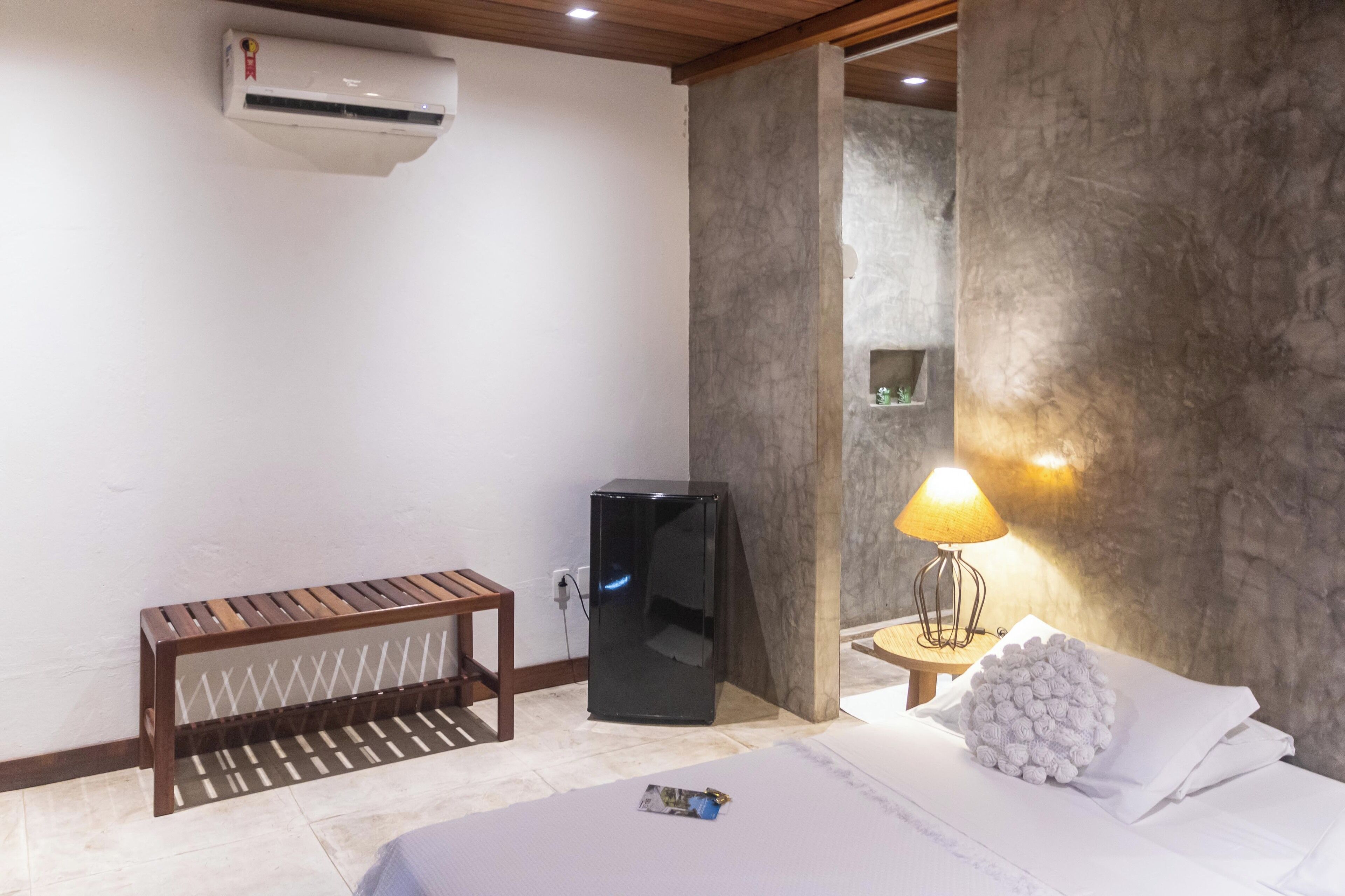 A hotel room with a bed, a small wooden bench, a black refrigerator, a wall-mounted air conditioner, and a lamp on a side table.