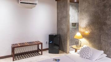 A hotel room with a bed, a small wooden bench, a black refrigerator, a wall-mounted air conditioner, and a lamp on a side table.