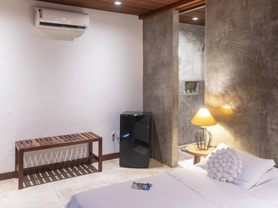 A hotel room with a bed, a small wooden bench, a black refrigerator, a wall-mounted air conditioner, and a lamp on a side table.