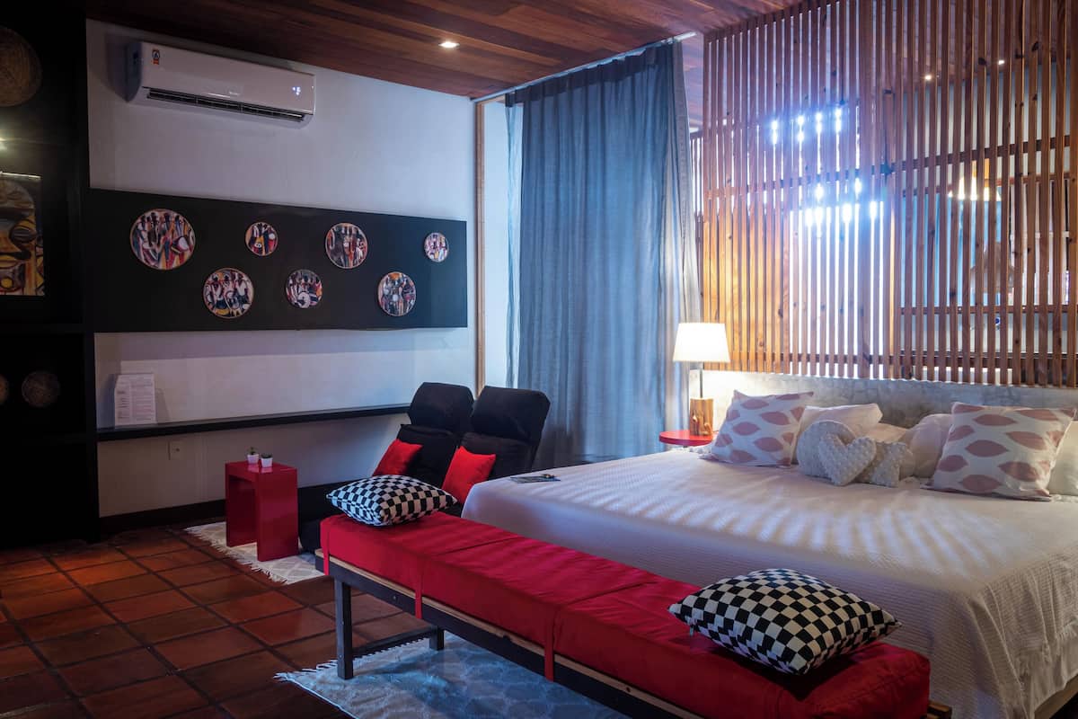 Habitación de hotel con una cama grande, sillas negras y rojas, un banco rojo, aire acondicionado de pared y una cortina.