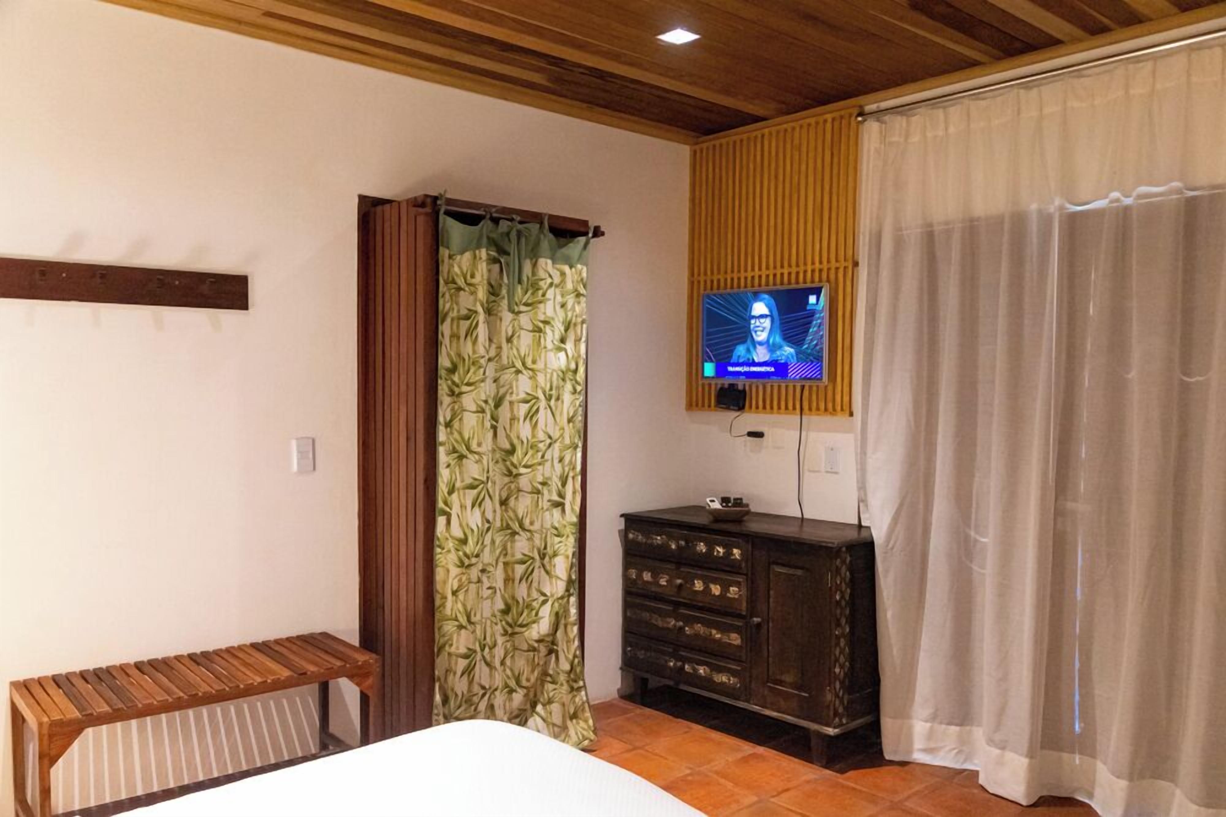 Un dormitorio con cama, banca de madera, televisor montado en la pared, cómoda y ventana con cortinas de bambú.