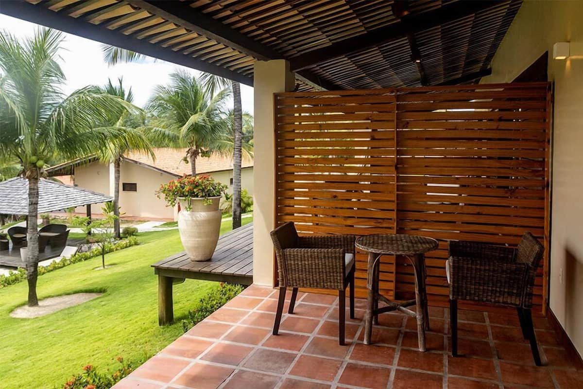 Un patio exterior con valla de madera, una mesa redonda, dos sillas de mimbre, una planta en maceta y un césped con palmeras.
