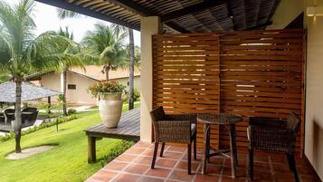 An outdoor patio with a wooden fence, a round table, two wicker chairs, a potted plant, and a lawn with palm trees.