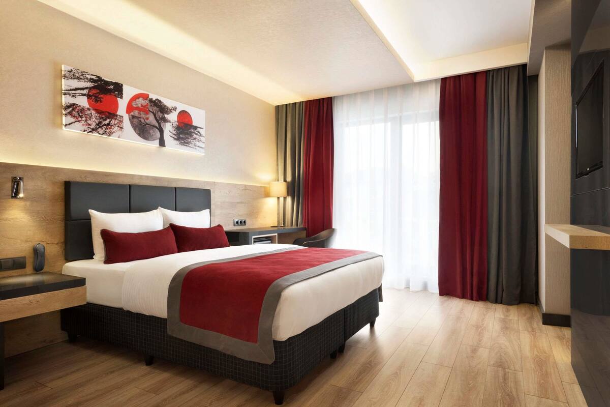 A modern hotel room with a large bed, a desk, a TV, and a painting on the wall.