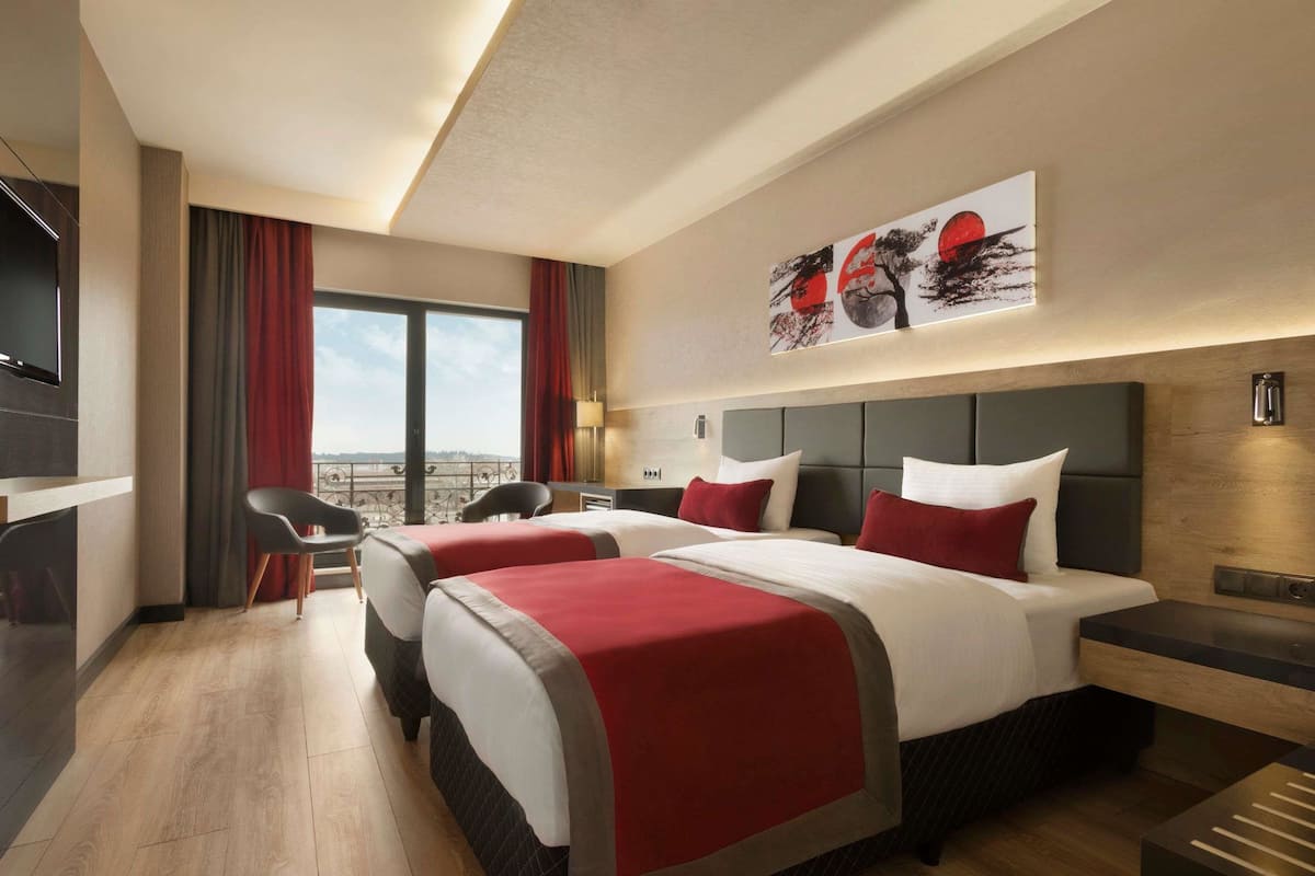 A hotel room with two beds, a desk, and a chair. There is a large window with a view of the city.