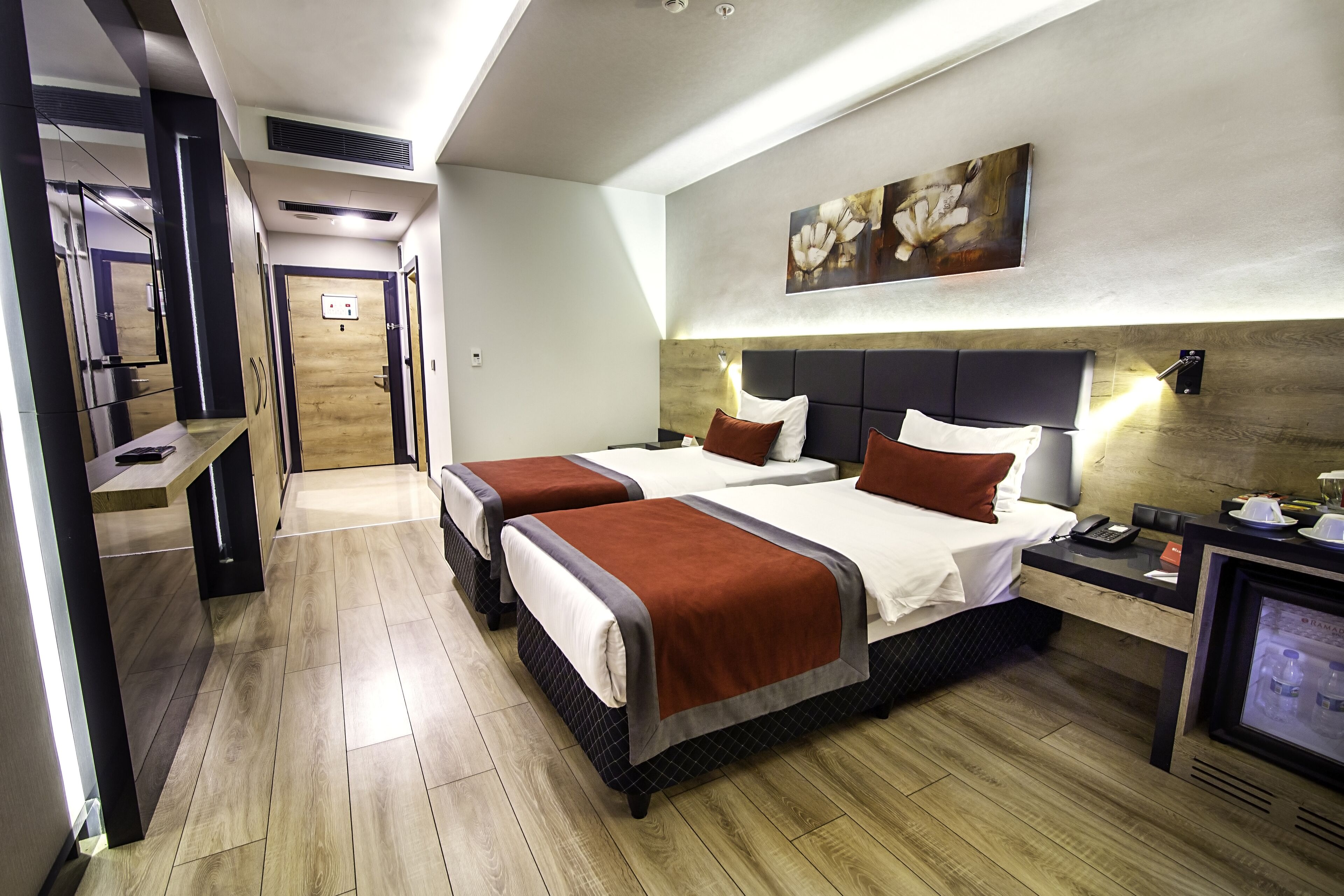 Una habitación de hotel con dos camas, piso de madera, cabecera con lámpara y mesita de noche con teléfono, además de una obra de arte colgada en la pared.