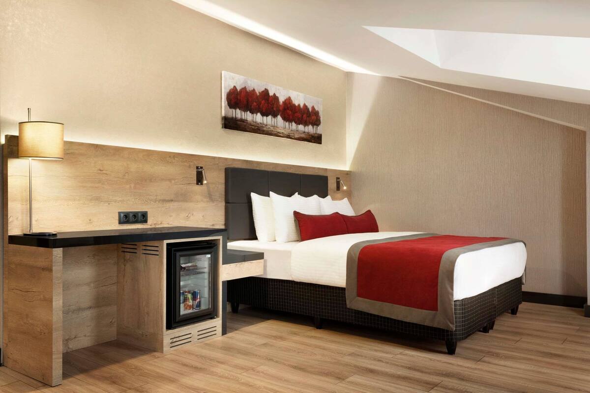 A modern hotel room with a bed, bedside table, lamp, and a painting on the wall.
