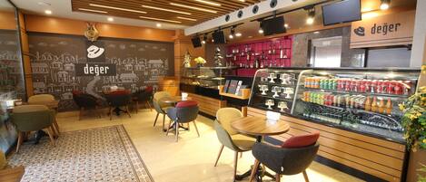 A cafe interior with wooden ceiling panels, a chalkboard wall with 'deger' written on it, a display case with beverages, and several chairs.