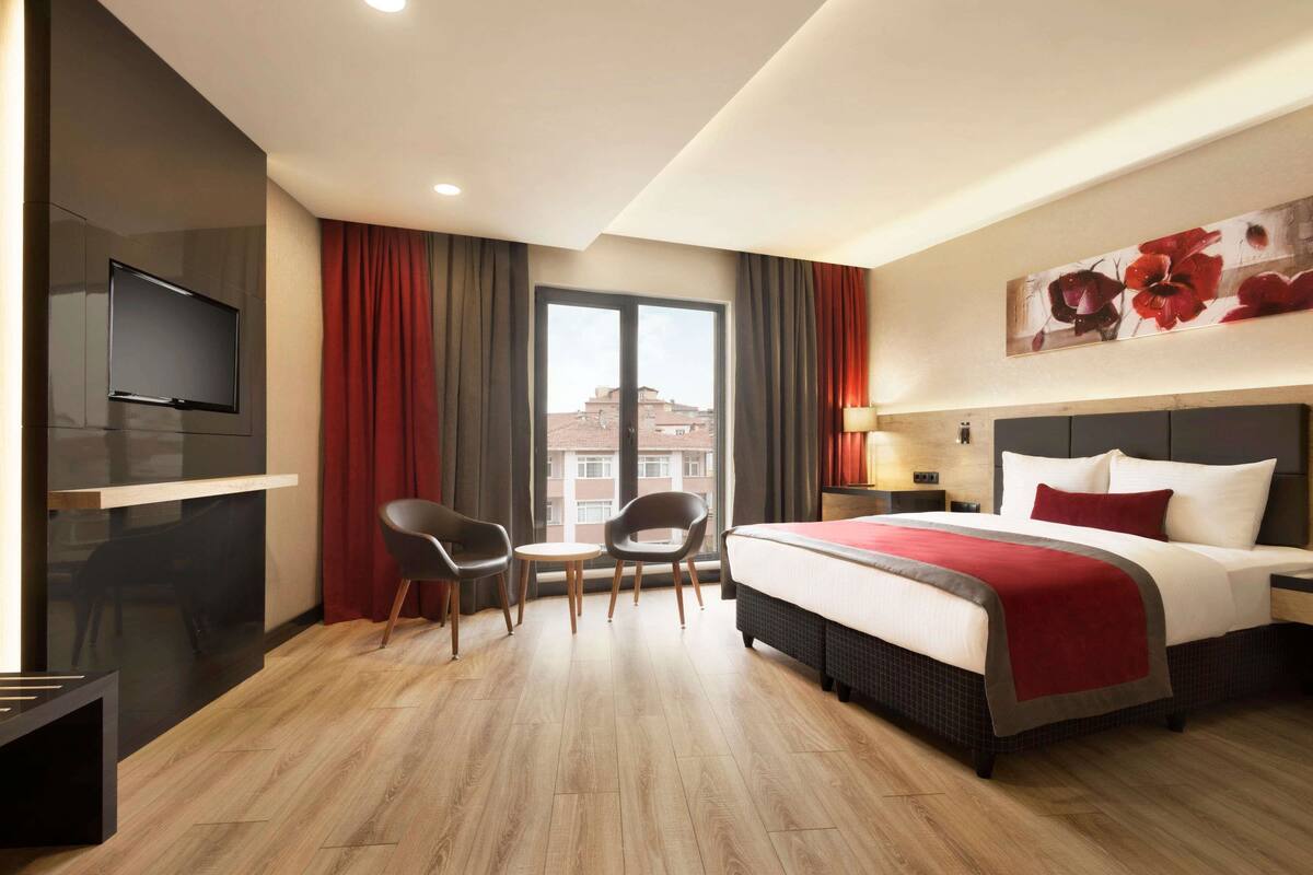 A hotel room with a bed, a TV mounted on the wall, a dining table with chairs, and a large window with curtains.