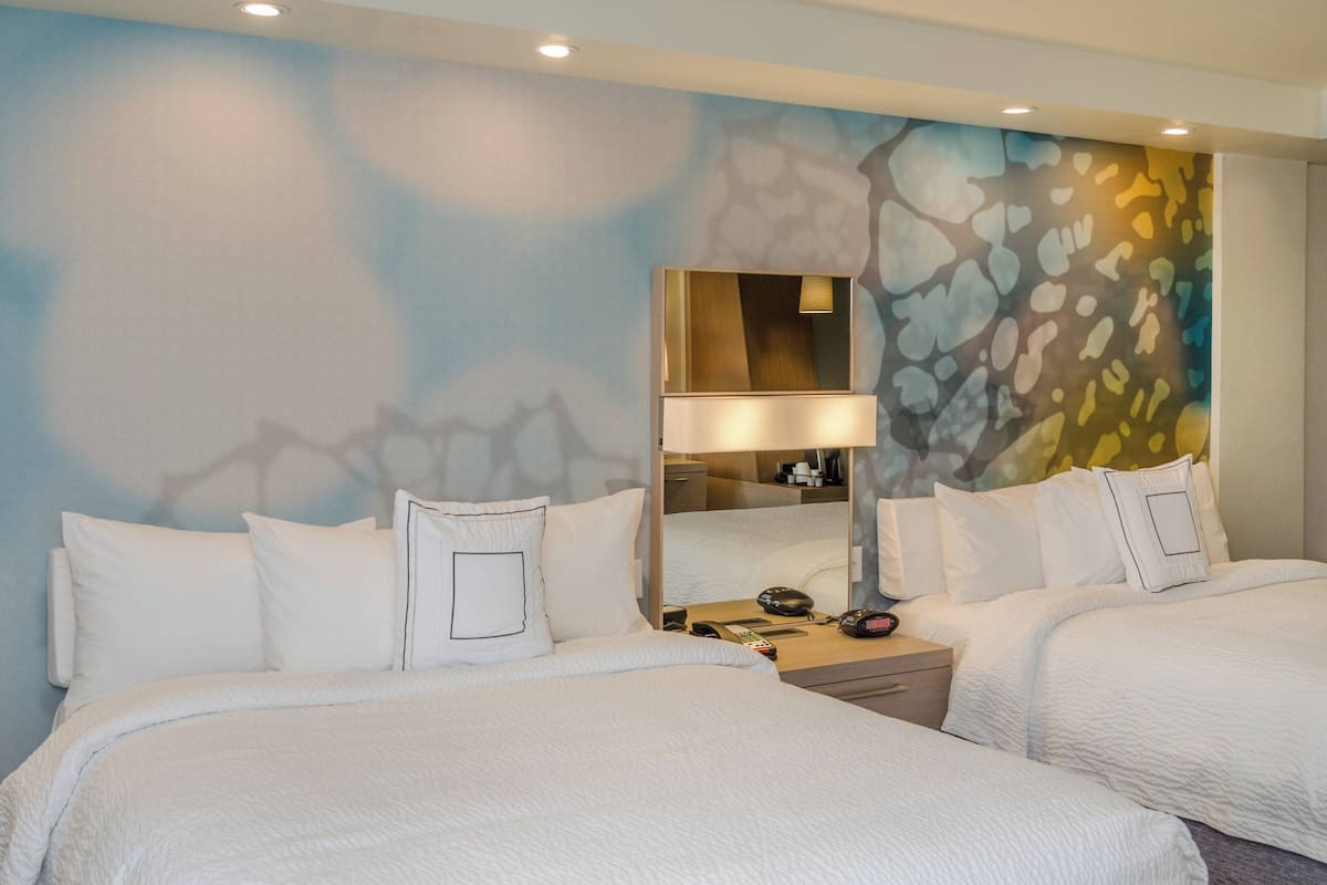 A hotel room with two beds, a nightstand with a lamp, a mirror, and a wall with a textured design.