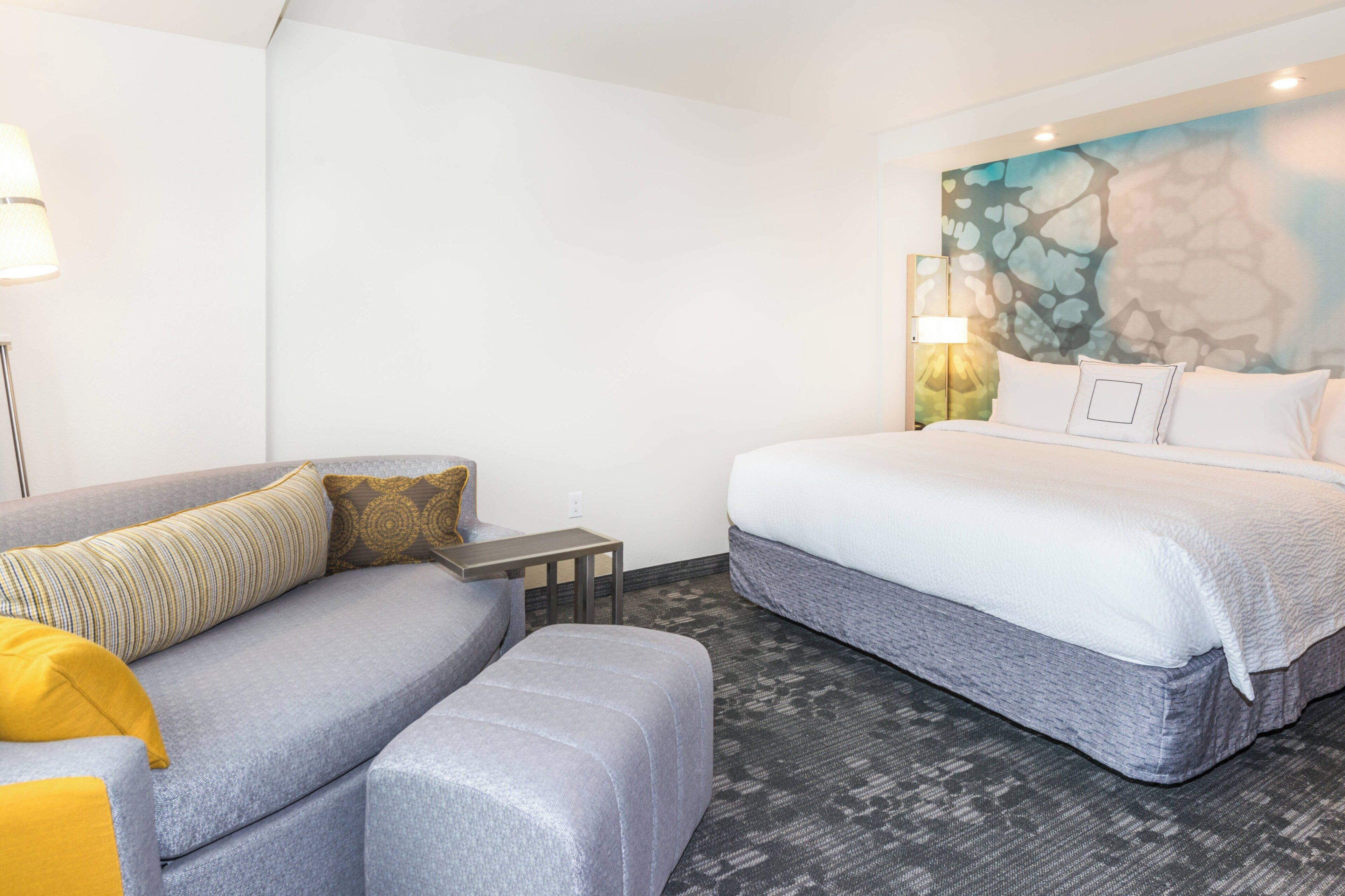 Una habitación de hotel con una cama, un sofá gris, una mesita pequeña y una pared con diseño texturizado.