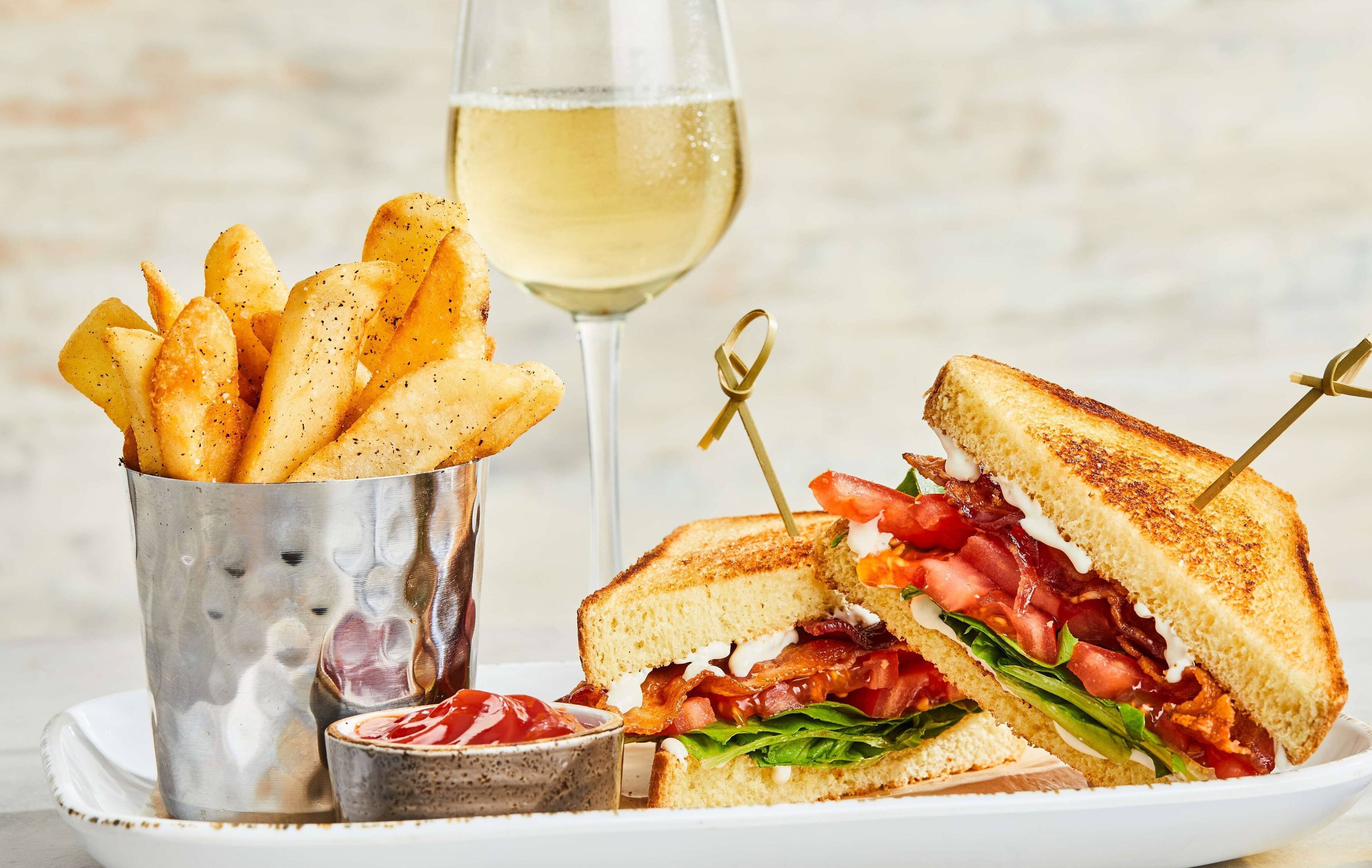 A sandwich with a toothpick, a glass of white wine, and a cup of seasoned fries.