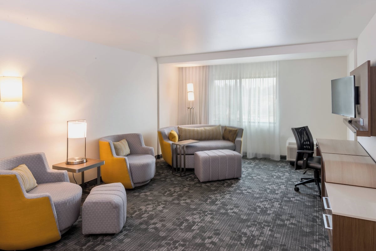 A hotel room with a grey sofa, yellow armchairs, a grey ottoman, a small table, a lamp, a TV, and a desk.