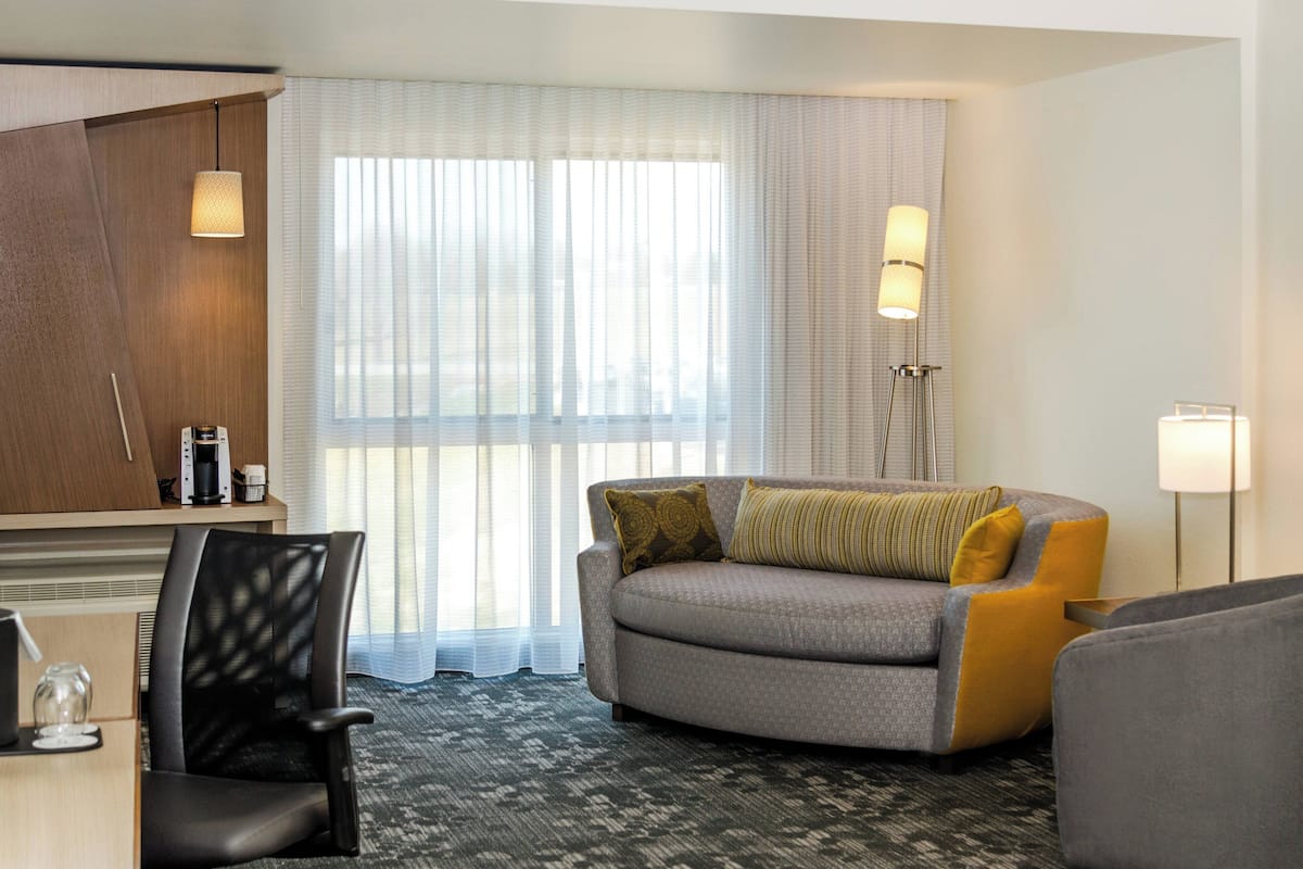A hotel room with a sofa, a chair, a desk, a lamp, and a large window with sheer curtains.