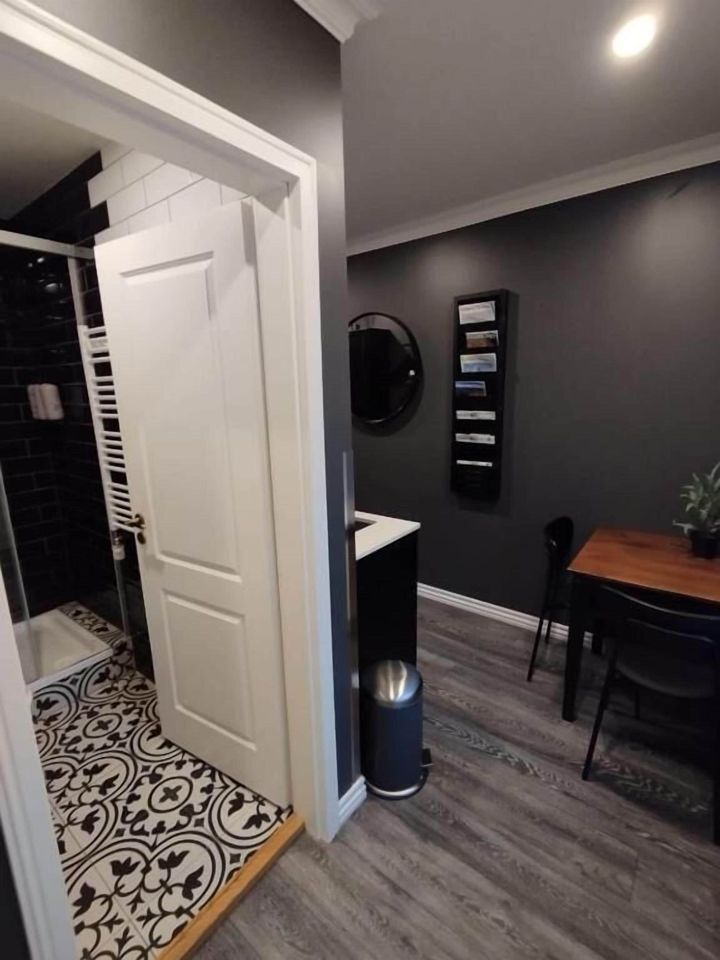 Un couloir avec une porte blanche, une poubelle noire, une table en bois avec des chaises et une étagère fixée au mur, sur laquelle se trouvent des papiers.